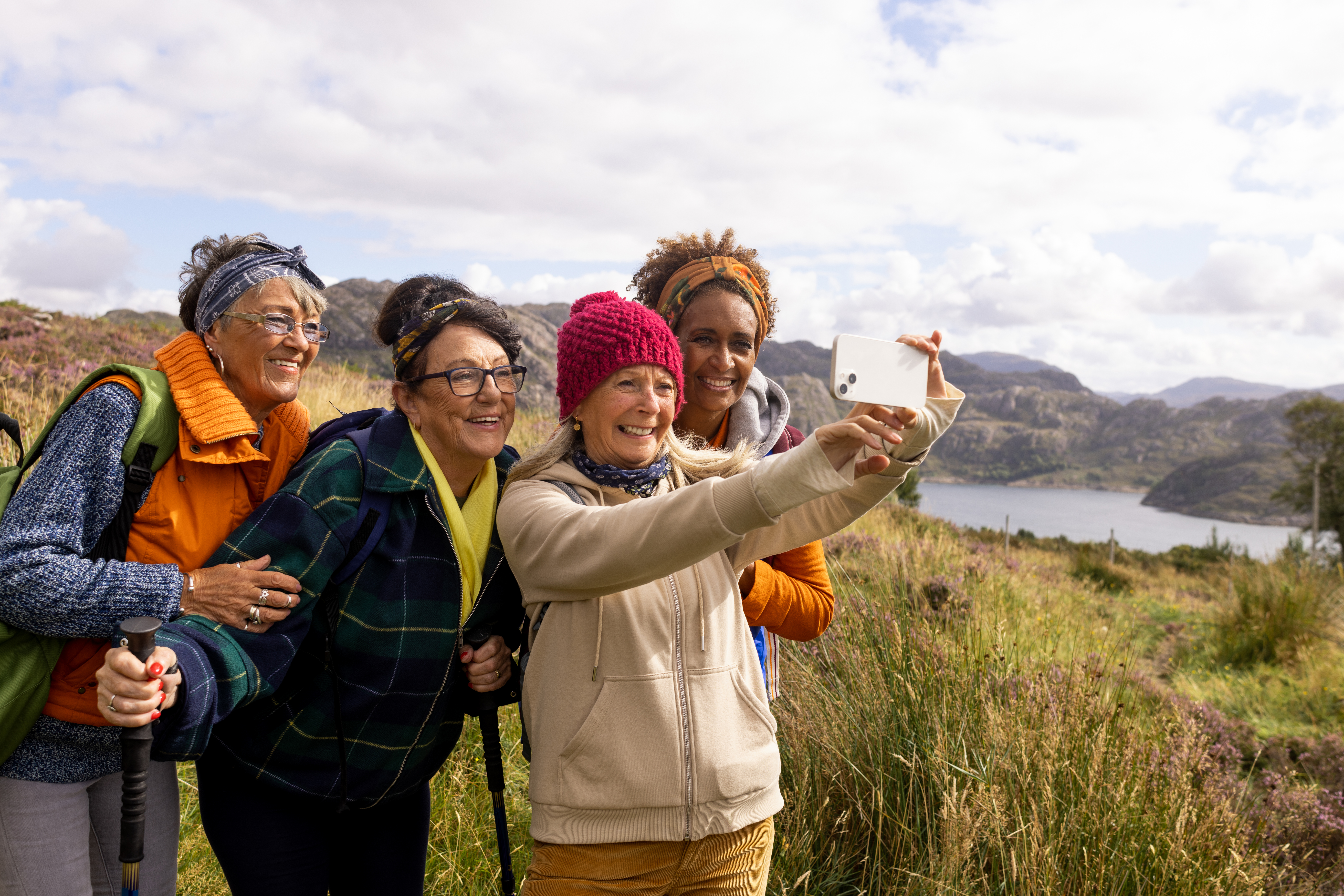 Front view of a small group of mature and senior female friends hiking up the mountain landscape surrounding Loch Torridon in the Northwest Highlands, Scotland. A senior woman is wearing a wooly hat and the rest are wearing bandanas, they are side by side taking a selfie on a smartphone.